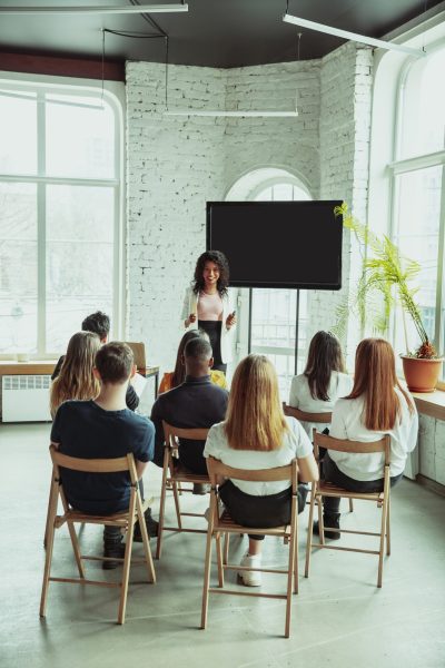 Female african-american speaker giving presentation in hall at workshop. Audience or conference hall. Rear view of participants in audience. Conference event, training. Education, diversity, inclusive concept.